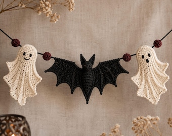 May include: A crochet garland featuring two off-white ghosts with embroidered facial features and a central black bat, all strung together on a dark cord interspersed with small, round, dark red textured beads.