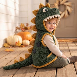Puede incluir: Un chaleco de dinosaurio tejido a ganchillo en color verde oscuro con capucha, dientes blancos, púas triangulares amarillas en la espalda y una cola larga. El chaleco incluye manchas circulares gris oscuro y bordes rematados en hilo amarillo.