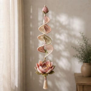 Op de afbeelding: Een hangende gehaakte mobiel met een spiraalvormige structuur in tinten crème, zachtroze en saliegroen. Het object wordt bekroond door een kleine gesloten bloemknop en eindigt in een grote, meerlaagse lotusbloem met een bungelend beige kwastje.