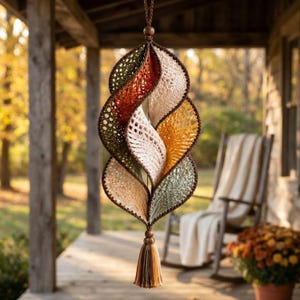 May include: A hanging spiral ornament crafted from crocheted yarn in shades of olive green, burnt orange, cream, and mustard yellow. The structure features a series of interconnected leaf-shaped segments with a dark brown border and a tassel at the base.