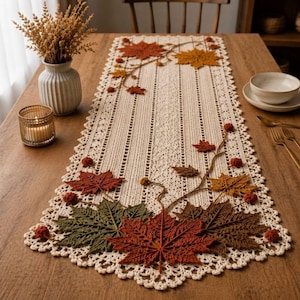 May include: A cream-colored crocheted table runner featuring an intricate lace border and a decorative pattern of three-dimensional fabric maple leaves in shades of red, orange, brown, and green, accented with small round pom-poms along the edges.