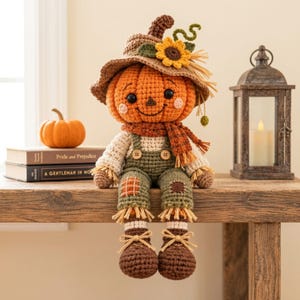 May include: A crocheted pumpkin-headed doll wearing a brown hat with a yellow sunflower, a plaid scarf, and green overalls with patches. The doll has black eyes, a stitched smile, and brown shoes with tan laces, sitting upright with dangling legs.