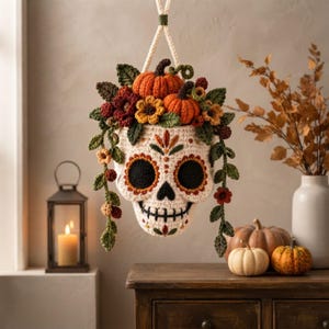Puede incluir: Una maceta colgante de ganchillo con forma de calavera, decorada con flores de lana de colores, hojas verdes y pequeñas calabazas naranjas. La calavera presenta cuencas oculares negras, una nariz negra y detalles de boca cosidos en lana crema, naranja y roja.