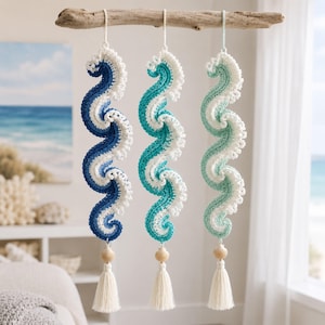 May include: Three hanging crochet wave decorations suspended from a piece of driftwood. Each wave features a gradient of blue, teal, or light green yarn with white foam-like edges. Each piece is finished with a small seashell and a white tassel.