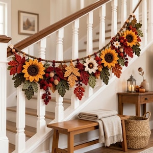 Op de afbeelding: Een decoratieve slinger met gehaakte zonnebloemen, esdoornbladeren en eikels in tinten geel, oranje, rood en groen. De slinger is gemaakt van een dik bruin touw en toont diverse botanische elementen met een herfstthema.