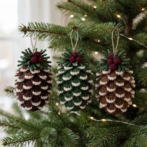 May include: Three crocheted pinecone ornaments hanging from a tree. Two are brown and one is green, all featuring white-tipped scales, dark red berries at the top, and green crocheted needles. Each ornament is suspended by a thin tan string loop.