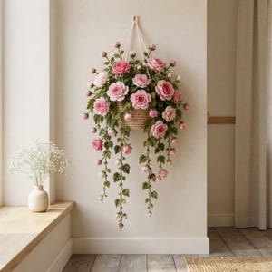 May include: A hanging woven basket filled with crocheted pink roses and green leaves. The arrangement features multiple blooming flowers and buds on long, trailing stems suspended by a white cord against a plain, light-colored wall surface.