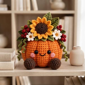 May include: A crocheted orange pumpkin figurine with a smiling face, black bead eyes, and pink cheeks. It is topped with a large yellow sunflower, small white flowers, green leaves, and clusters of red berries, with two brown feet at the base.