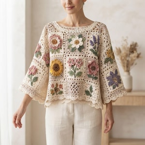 May include: A cream-colored crochet sweater featuring a square-patterned design with various three-dimensional floral motifs in shades of pink, yellow, green, and purple. The garment has wide sleeves, a wide neckline, and a scalloped hem edge.