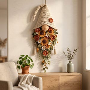Puede incluir: Una decoración de pared de ganchillo con forma de gnomo que presenta un sombrero puntiagudo de color crema, una nariz redonda y una larga barba compuesta por girasoles, margaritas y hojas verdes tejidas en tonos naranja, amarillo y marrón.