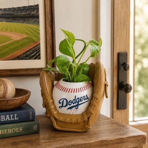 May include: A ceramic planter shaped like a baseball with red stitching and the blue Dodgers logo, nestled inside a tan leather baseball glove. A green leafy plant grows from the top of the baseball-shaped container, creating a sports-themed display.