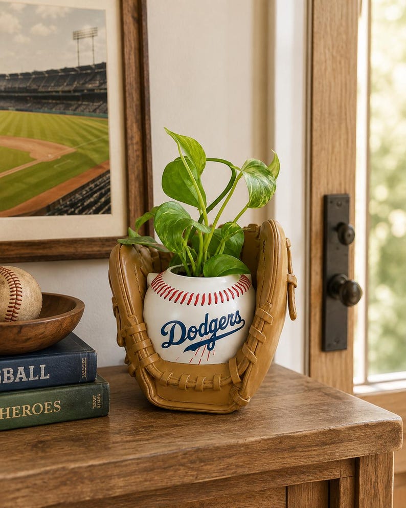 May include: A ceramic planter shaped like a baseball with red stitching and the blue Dodgers logo, nestled inside a tan leather baseball glove. A green leafy plant grows from the top of the baseball-shaped container, creating a sports-themed display.