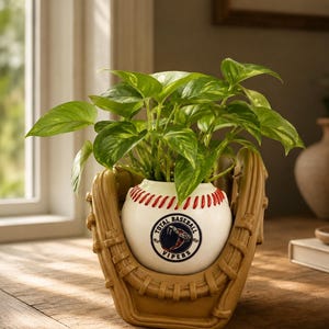 May include: A tan baseball glove-shaped planter holding a white ceramic baseball with red stitching. The baseball features a circular logo with a snake graphic and the text Total Baseball Vipers. A green leafy plant grows from the top of the ball.