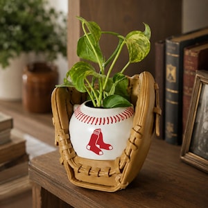 Peut inclure: Un gant de baseball en cuir beige tenant un pot en céramique blanc en forme de balle de baseball. Le pot présente des coutures rouges et un motif rouge représentant deux chaussettes. Une plante verte pousse au sommet du pot en céramique.