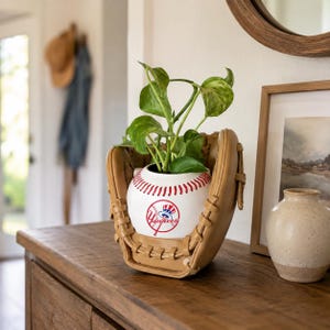 May include: A tan baseball glove holding a white ceramic planter shaped like a baseball with red stitching. The planter features a red, white, and blue New York Yankees logo with a top hat graphic. A green leafy plant grows from the baseball planter.