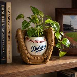 May include: A white ceramic planter shaped like a baseball with the blue Dodgers logo and red stitching, nestled inside a tan baseball glove. A green leafy plant grows out of the top of the baseball planter, with vines trailing over the side.