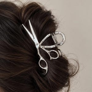 May include: A silver-toned metal hair claw clip shaped like a pair of scissors. The hair accessory features a polished finish and a spring-loaded mechanism, securing a dark brown hair bun in place. The design mimics the form of professional shears.