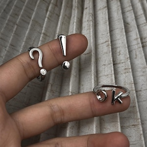 Op de afbeelding: Een set zilverkleurige metalen ringen met leestekens en letters. Eén ring heeft de vorm van een vraagteken, een andere van een uitroepteken en een derde ring bevat de letters O en K in een open ringontwerp met een glanzende afwerking.