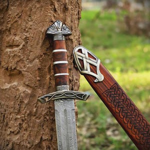 May include: A sword with a patterned Damascus steel blade, a dark wood handle with silver-coloured metal bands, and a matching wooden scabbard featuring intricate carved knotwork patterns and a decorative silver-coloured metal chape at the top.
