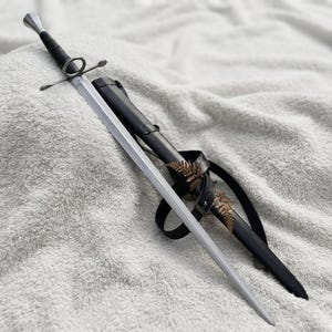 Può includere: Una spada in metallo color argento con impugnatura avvolta in nero, guardia circolare e traversa dritta. La spada è accompagnata da un fodero in pelle nera con cinturino, impreziosito da una piccola foglia di felce essiccata marrone.