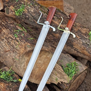 May include: Two swords with polished steel blades and dark reddish-brown wooden handles. Each handle features a row of small metal rivets. One sword has a silver-coloured metal crossguard, while the other has a gold-coloured metal crossguard.