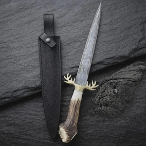 May include: A fixed-blade knife with a patterned Damascus steel blade, a brass crossguard shaped like deer antlers, and a textured brown antler handle. The knife is accompanied by a black leather sheath with a snap button closure.