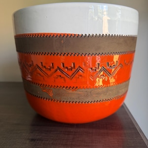 May include: A cylindrical ceramic planter featuring a white upper rim, a vibrant orange lower section, and two horizontal bands of matte brown texture. The orange area is decorated with a repeating geometric zigzag pattern carved into the surface.