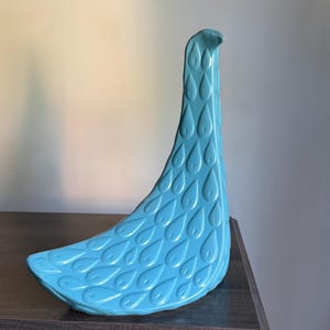 May include: A turquoise ceramic decorative bird figurine with a long neck and a curved body. The bird's body is adorned with a pattern of teardrop-shaped indentations. The figurine is displayed on a dark wooden surface.