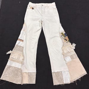 May include: Off-white denim jeans featuring wide, flared legs with patchwork inserts. The side panels include various textured fabrics in cream, beige, and tan, accented with lace, fringe, and frayed edges. The jeans have a standard button fly.