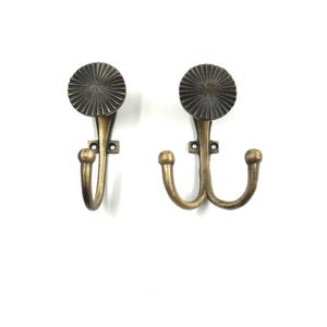 Peut inclure: Deux patères en métal couleur laiton antique. Le crochet de gauche présente un bras incurvé unique avec une extrémité sphérique, tandis que celui de droite possède un double bras incurvé. Les deux sont surmontés d'un disque circulaire.