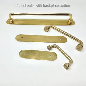 May include: A collection of polished brass hardware including fluted pulls and rectangular backplates with rounded ends. The text fluted pulls with backplate option is visible at the top. These metal components feature a smooth, reflective finish.