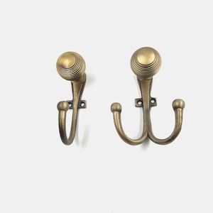 Peut inclure: Deux patères murales en laiton antique avec une finition métallique. Le crochet de gauche présente un bras incurvé unique et un sommet sphérique strié. Le crochet de droite a un design à double bras incurvé avec un sommet sphérique strié assorti.