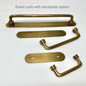 Könnte beinhalten: Eine Sammlung von Schrankbeschlägen aus messingfarbenem Metall, bestehend aus geriffelten Griffen und passenden rechteckigen Rückplatten mit abgerundeten Enden. Der Text "fluted pulls with backplate option" ist oben zu sehen.