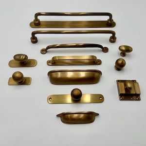 Op de afbeelding: Een collectie kastbeslag van antiek messing, inclusief diverse ladehandgrepen, komgrepen, knoppen en een grendel. De set heeft een geborstelde metalen afwerking met gladde, afgeronde randen en zichtbare schroefgaten op de montageplaten.