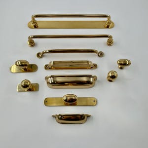 May include: A collection of polished brass hardware including various drawer pulls, cabinet handles, and knobs. The set features long bar pulls with rectangular backplates, curved cup pulls, and round knobs with both circular and rectangular backplates.