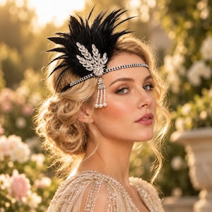May include: A decorative headpiece featuring a black feather plume attached to a silver-toned band encrusted with clear rhinestones. The design includes a central leaf-shaped rhinestone ornament with a dangling tassel of small white faux pearls.