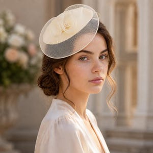 Peut inclure: Un bibi de couleur crème doté d'une base circulaire, d'une superposition en maille transparente et d'un accent floral central orné de petites perles blanches. La coiffe est structurée avec un bord en tissu rigide et des détails en filet.