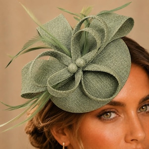 May include: A sage green fascinator hat featuring a circular base with a textured woven fabric finish. The headpiece is adorned with large, structured loops of the same material and delicate, thin feathers extending outward from the center.