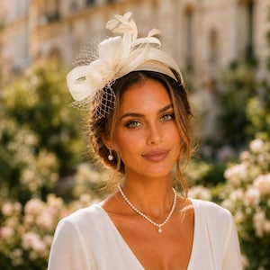 Può includere: Un fascinator color avorio con anelli strutturati, piume delicate e un velo a rete fine. L'accessorio è abbinato a una semplice collana di perle e a un orecchino con perla pendente, su un capo bianco con scollo a V.
