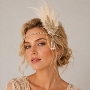 May include: A gold-toned headband featuring a thin, rhinestone-encrusted band, an upright cream-colored feather plume, a gold leaf-shaped embellishment with clear crystals, and a dangling fringe of small white pearls on one side.