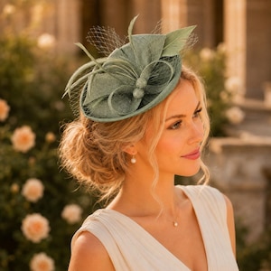 Può includere: Un fascinator verde salvia caratterizzato da una base circolare strutturata, decorativi passanti in tessuto e un delicato velo a rete. L'accessorio è ornato da piume sottili e dritte che aggiungono altezza e consistenza al design.