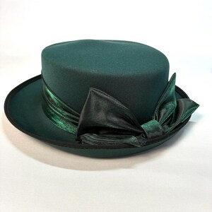 May include: A dark forest green hat with a flat top and a wide brim. The hat features a dark green satin ribbon band wrapped around the base, tied into a large, prominent bow on the side. The brim edge is finished with a thin black trim.