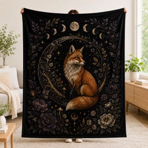 Peut inclure: Un plaid rectangulaire noir orné d'une illustration centrale représentant un renard roux assis sur un croissant de lune. Le motif comprend un cycle lunaire sur le haut, entouré de motifs floraux complexes, de petits champignons et d'étoiles.