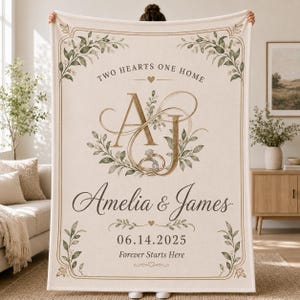 May include: A cream-colored throw blanket featuring a decorative border with green leaves. The text reads Two Hearts One Home, Amelia & James, 06.14.2025, and Forever Starts Here. Large gold letters AJ are centered with a ring illustration.