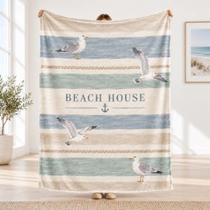 Könnte beinhalten: Eine weiche Wohndecke mit horizontalen Streifen in Beige, Hellblau und Weiß. Das Design zeigt vier Möwen in verschiedenen Flug- und Standpositionen, seilartige Akzente sowie den Schriftzug BEACH HOUSE mit einem Anker-Symbol.