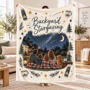 Puede incluir: Una manta de forro polar color crema con el texto "Backyard Stargazing" en letra azul oscuro. La ilustración central muestra a una familia y un perro sentados sobre una manta a cuadros bajo un cielo estrellado con una luna creciente.