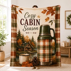 Könnte beinhalten: Eine rechteckige Wohndecke mit einer Waldhütte, einer Tasse, einem Tannenzapfen und einer großen Thermoskanne. Das Design zeigt den Text "Cozy CABIN Season" mit Herbstblättern und einem karierten Rand in Grün-, Orange- und Brauntönen.