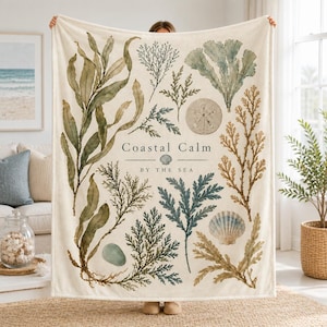 May include: A cream-coloured throw blanket featuring a botanical print of various seaweeds, a sand dollar, and a seashell. The text "Coastal Calm BY THE SEA" is printed in the centre in a dark grey serif font, surrounded by green and brown illustrations.