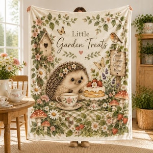 Puede incluir: Una manta de color crema con una ilustración central de un erizo con una taza de té y un pastel. El texto dice Little Garden Treats y Good Things Grow Here. El diseño incluye una pajarera, mariposas, fresas, setas y enredaderas florales.