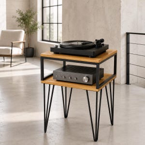 May include: A two-tier side table with a black metal frame and light wood shelves. The top shelf holds a black record player, while the bottom shelf supports a black audio amplifier with silver knobs. The table features four black hairpin legs.